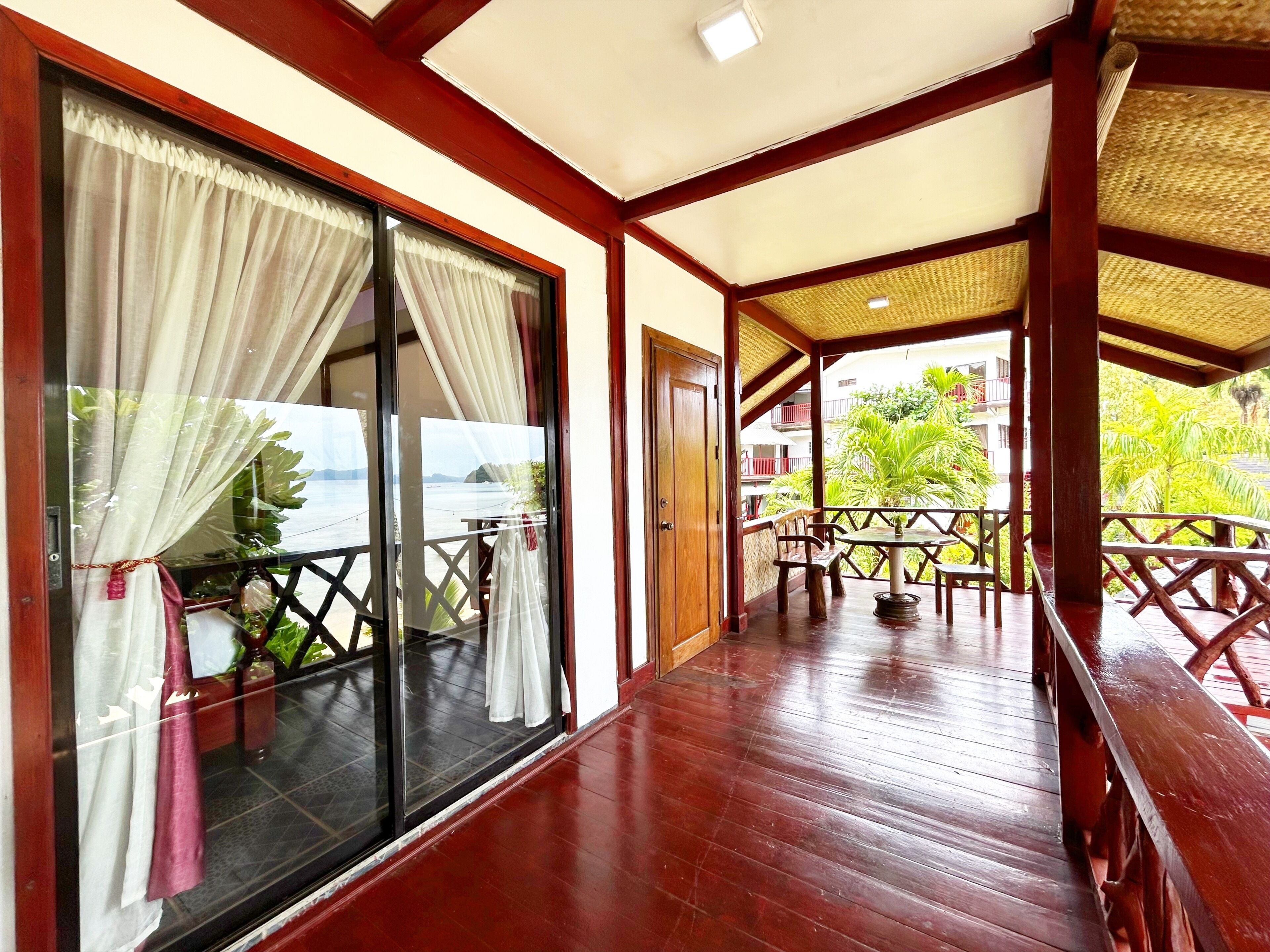 A wooden deck with a glass door leading to a balcony, a table, chairs, and a view of greenery.