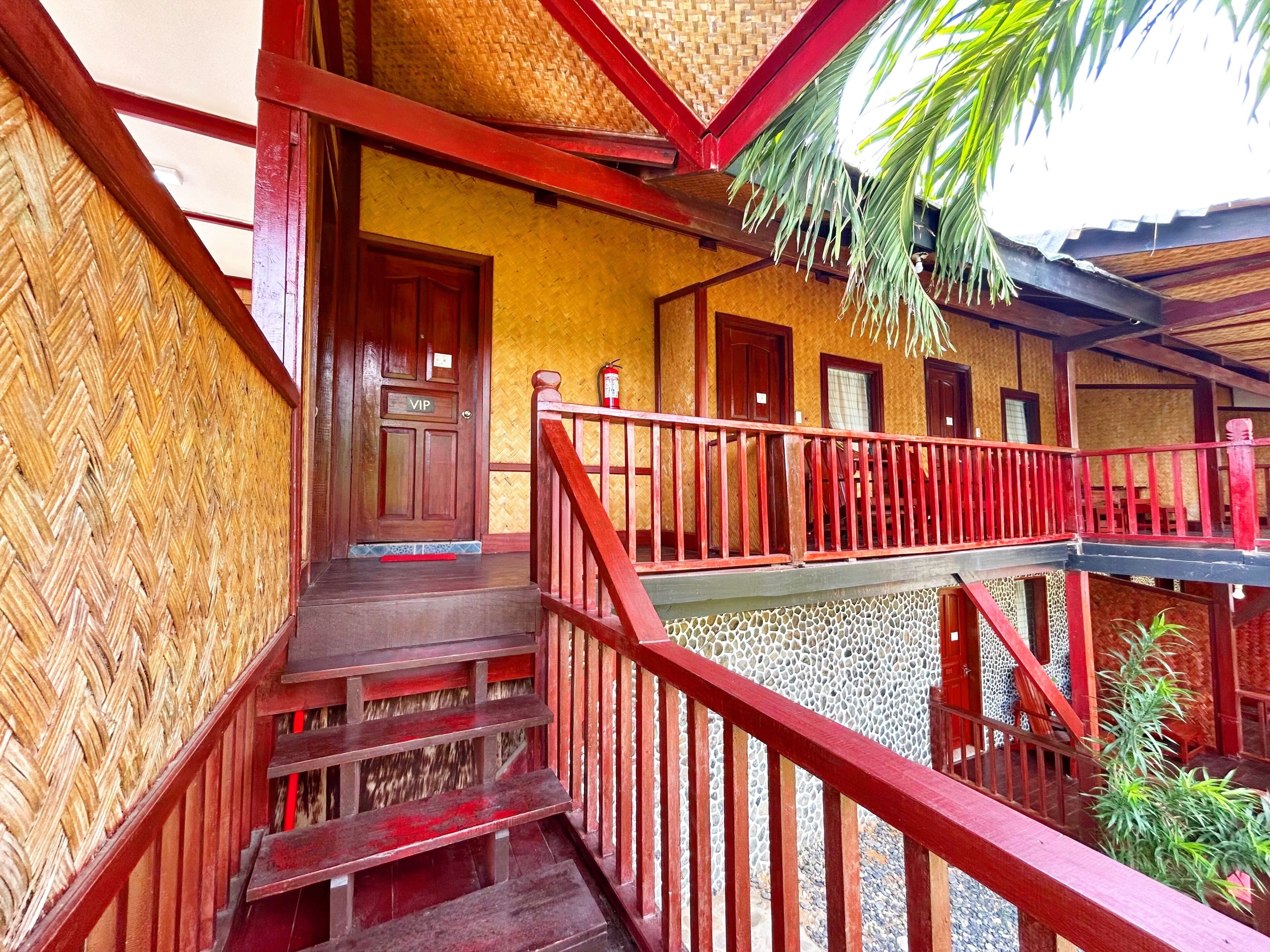 Uma escadaria de madeira com corrimão vermelho leva a uma varanda com vista para um edifício com telhado de palha.