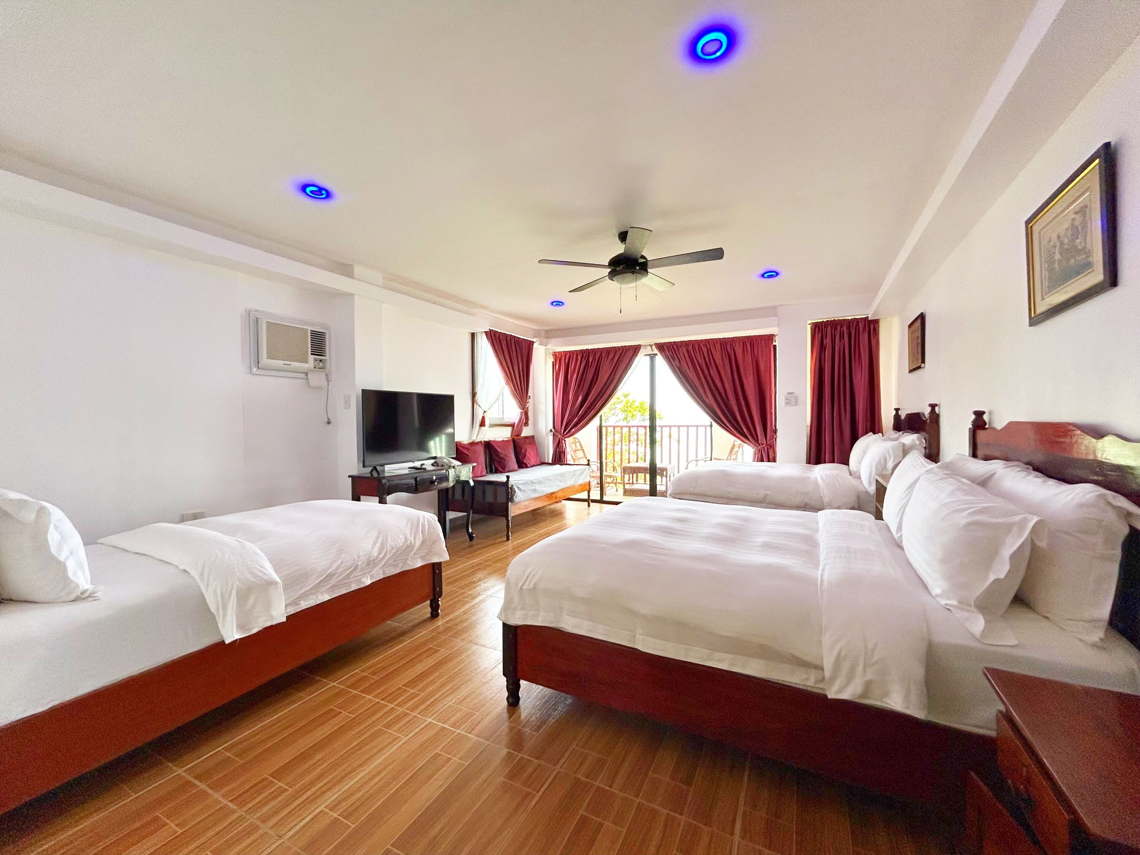 A hotel room with two beds, a TV, a ceiling fan, and a balcony with red curtains.