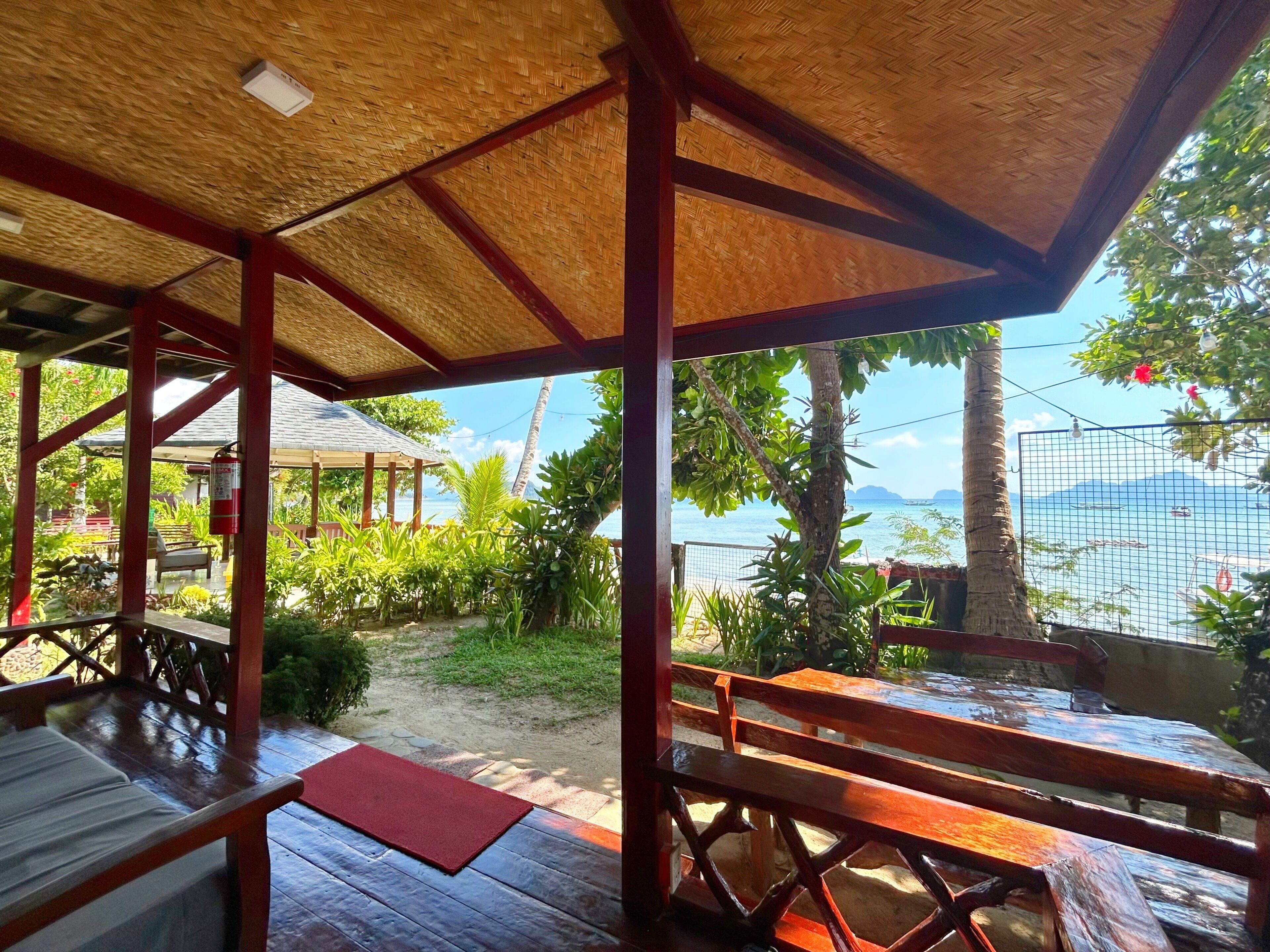 A covered outdoor area with wooden benches and tables, a thatched roof, and a view of the sea.