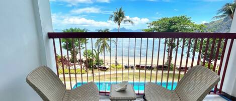A balcony with two wicker chairs and a small table, overlooking a pool and beach with palm trees and mountains in the distance.
