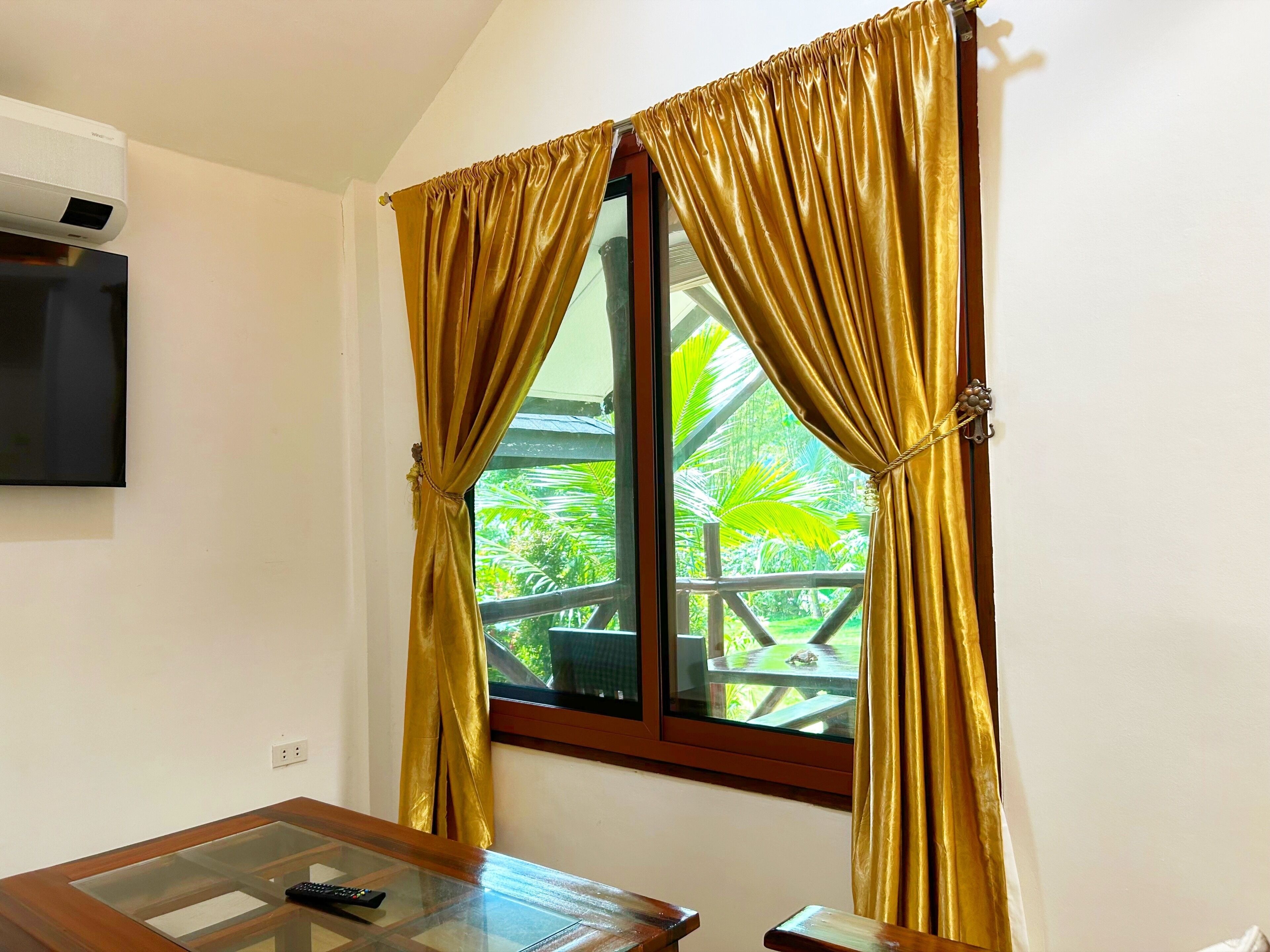 A room with a glass-top table, a remote control, and a window with golden curtains.