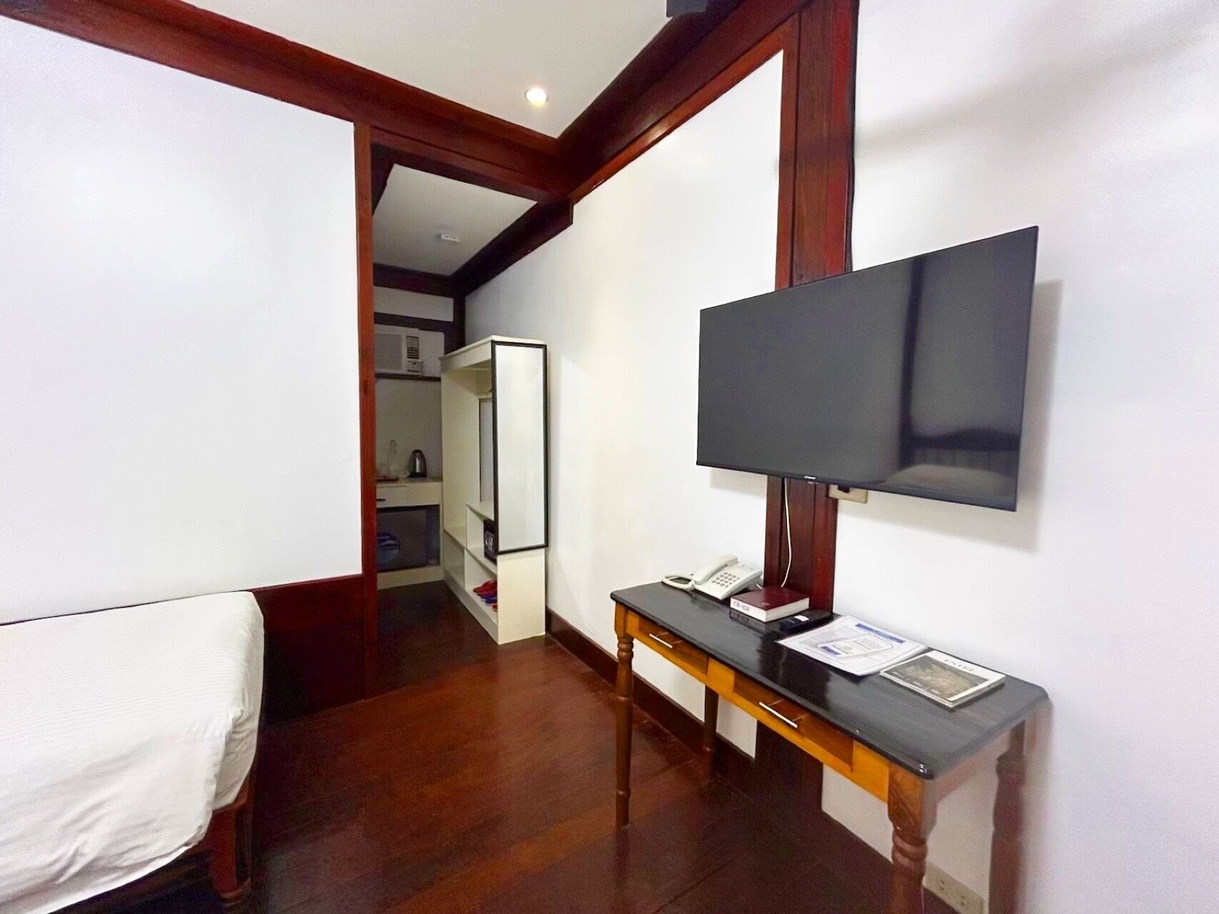 A hotel room with a bed, a desk with a telephone, a flat-screen TV mounted on the wall, and a wooden floor.
