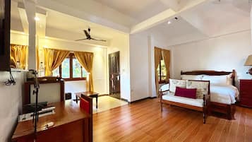 A spacious room with a wooden floor, a bed with a headboard, a desk with a chair, a ceiling fan, and a window with curtains.