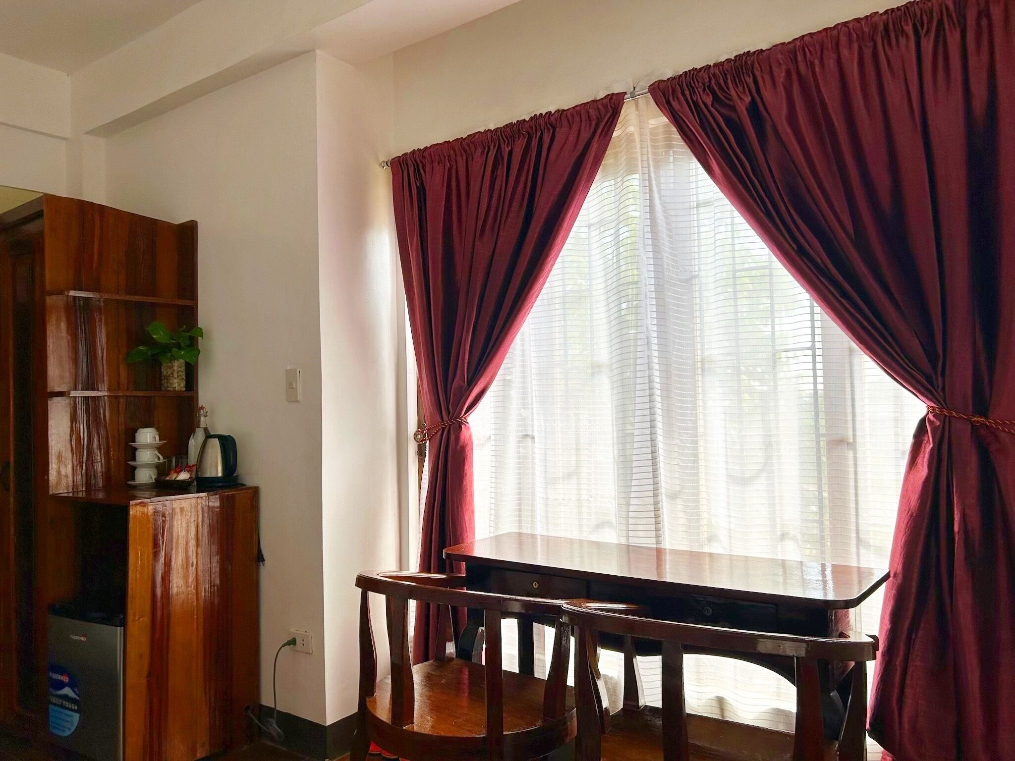 A room with a wooden desk, two chairs, a cabinet with a plant, and a window with red curtains.