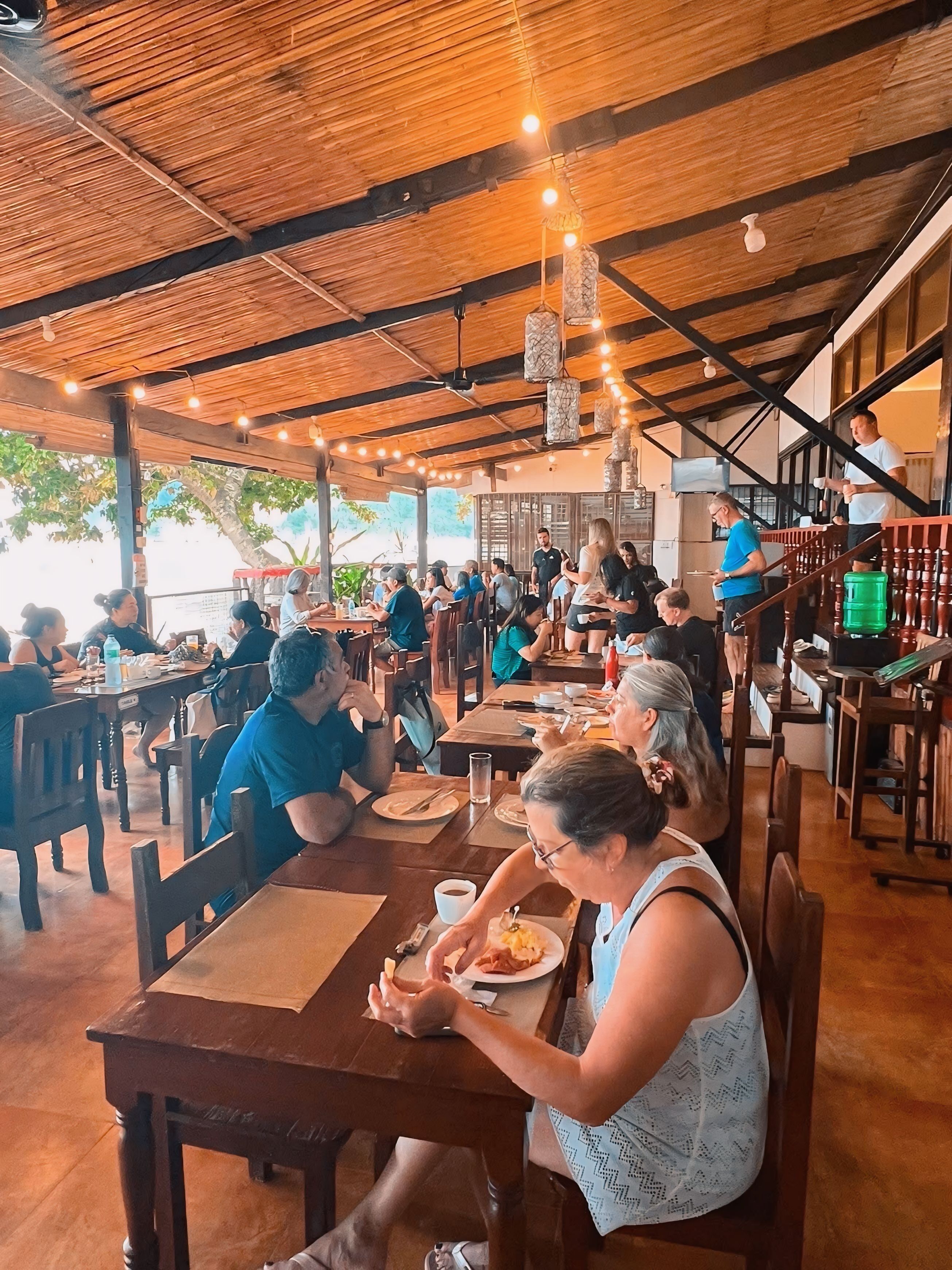Un restaurant avec des tables et des chaises en bois, des lampes suspendues et un toit de chaume.