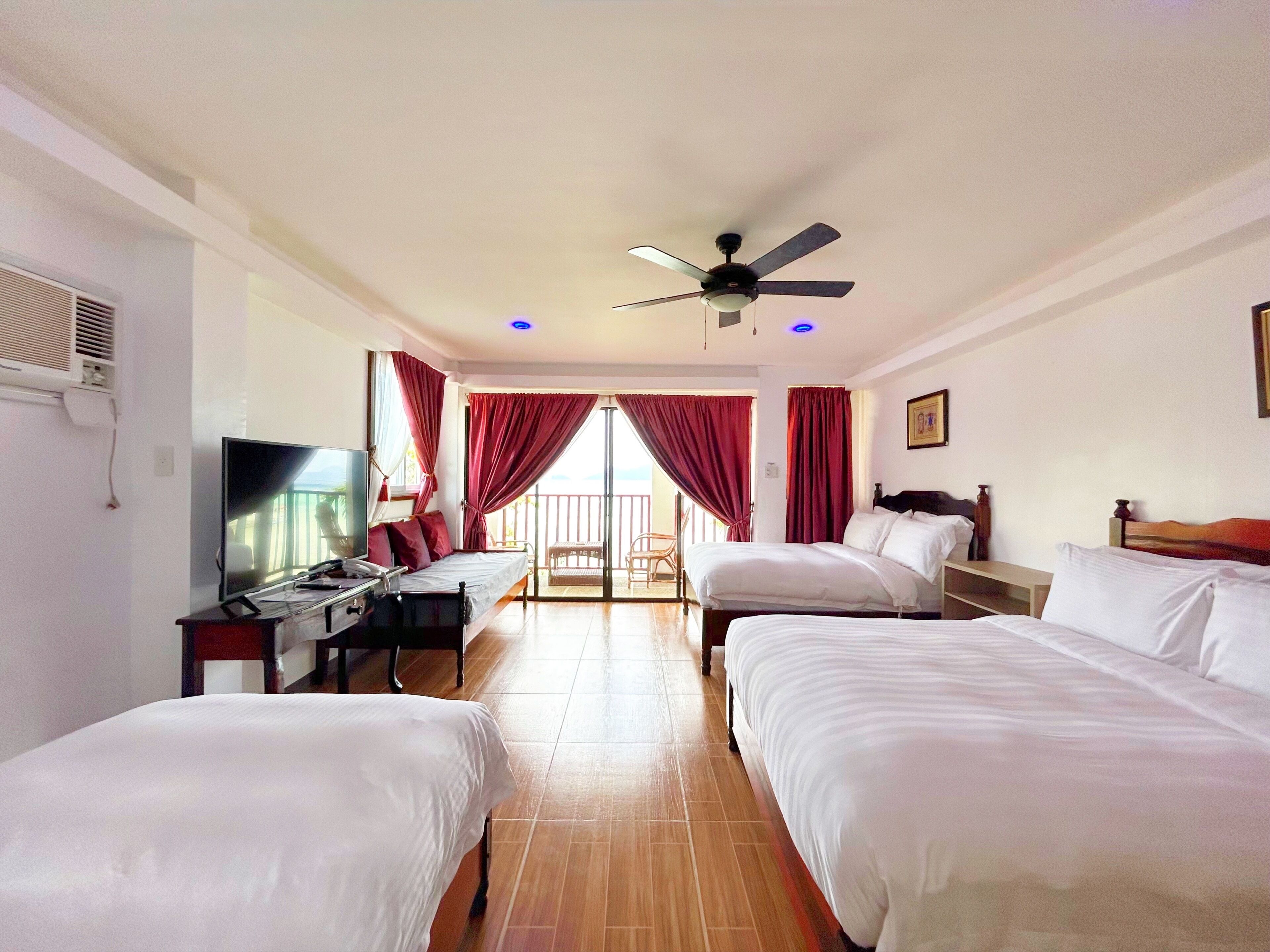 Quarto de hotel com duas camas, uma mesa de escritório com um computador, uma televisão e um ventilador de teto.