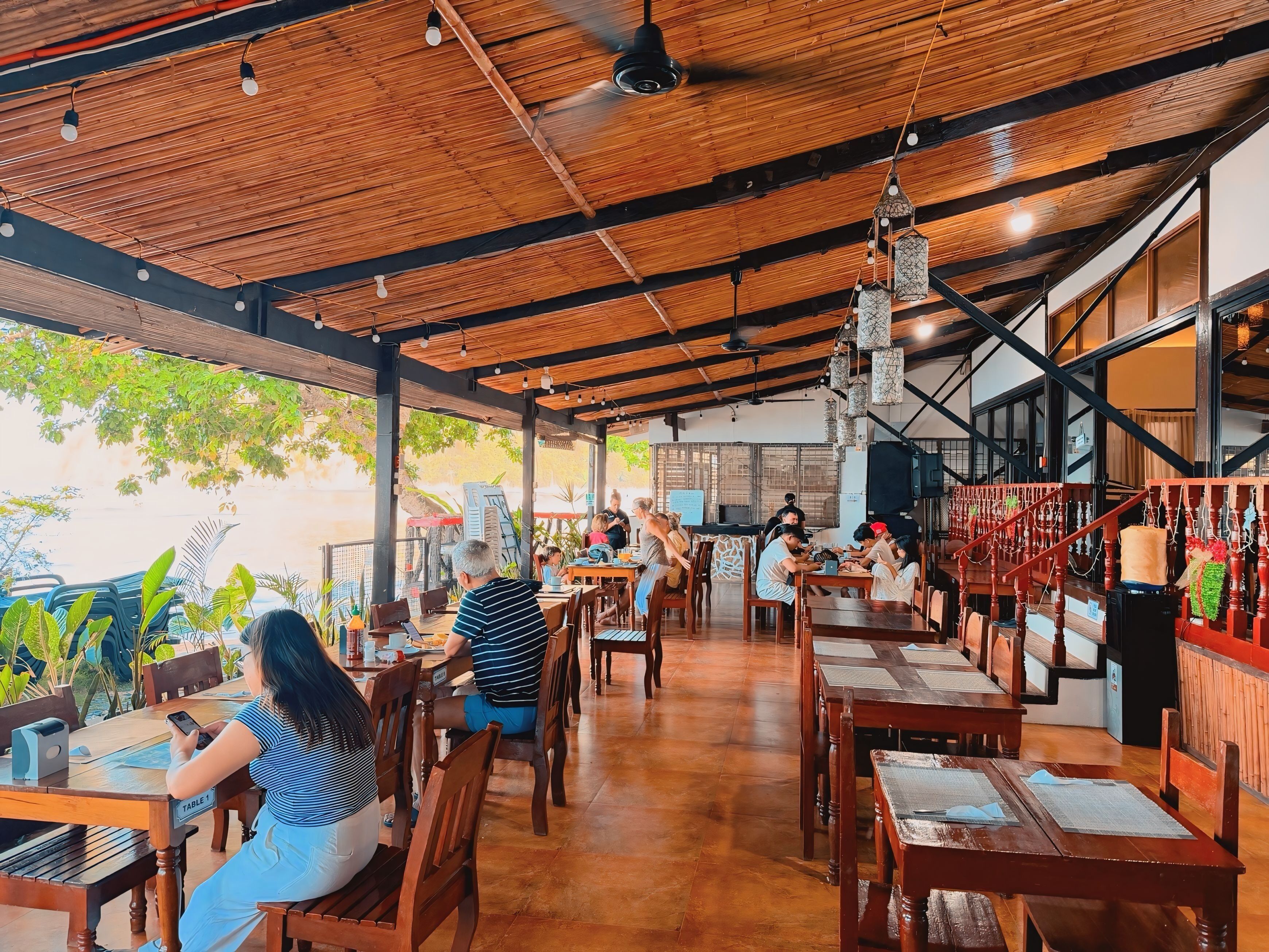 Ein Restaurant mit Holztischen und -stühlen, einer hohen Decke mit freiliegenden Balken und hängenden Laternen.