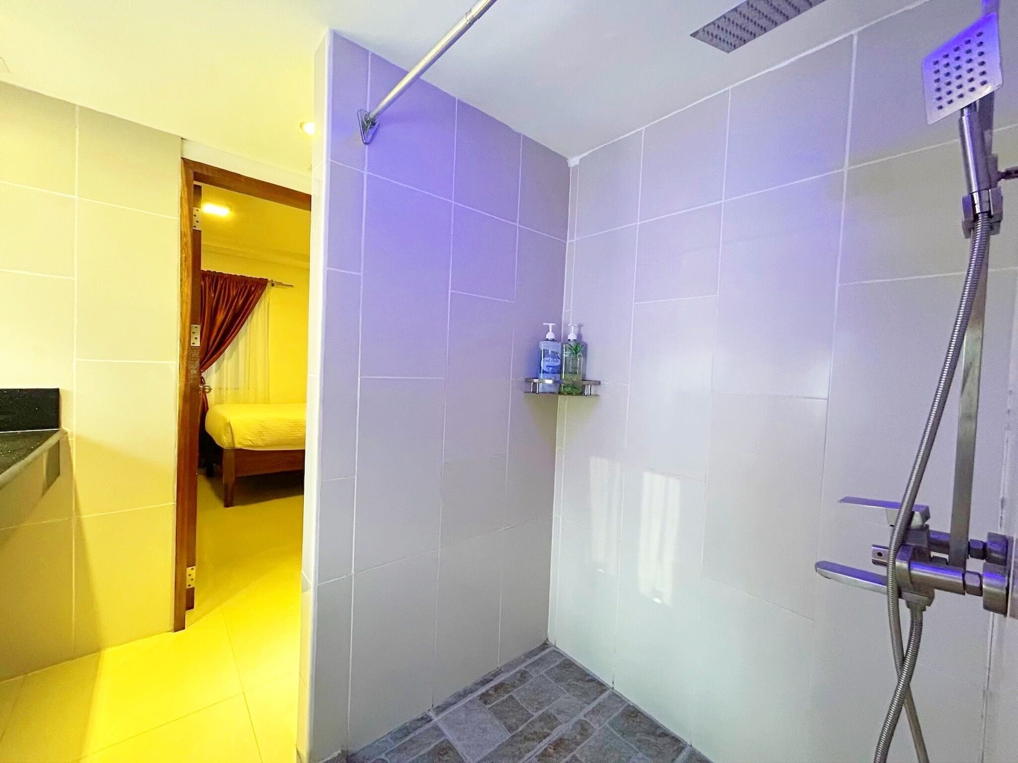A bathroom with a shower, a toilet, and a sink. There is a bed visible through an open door.