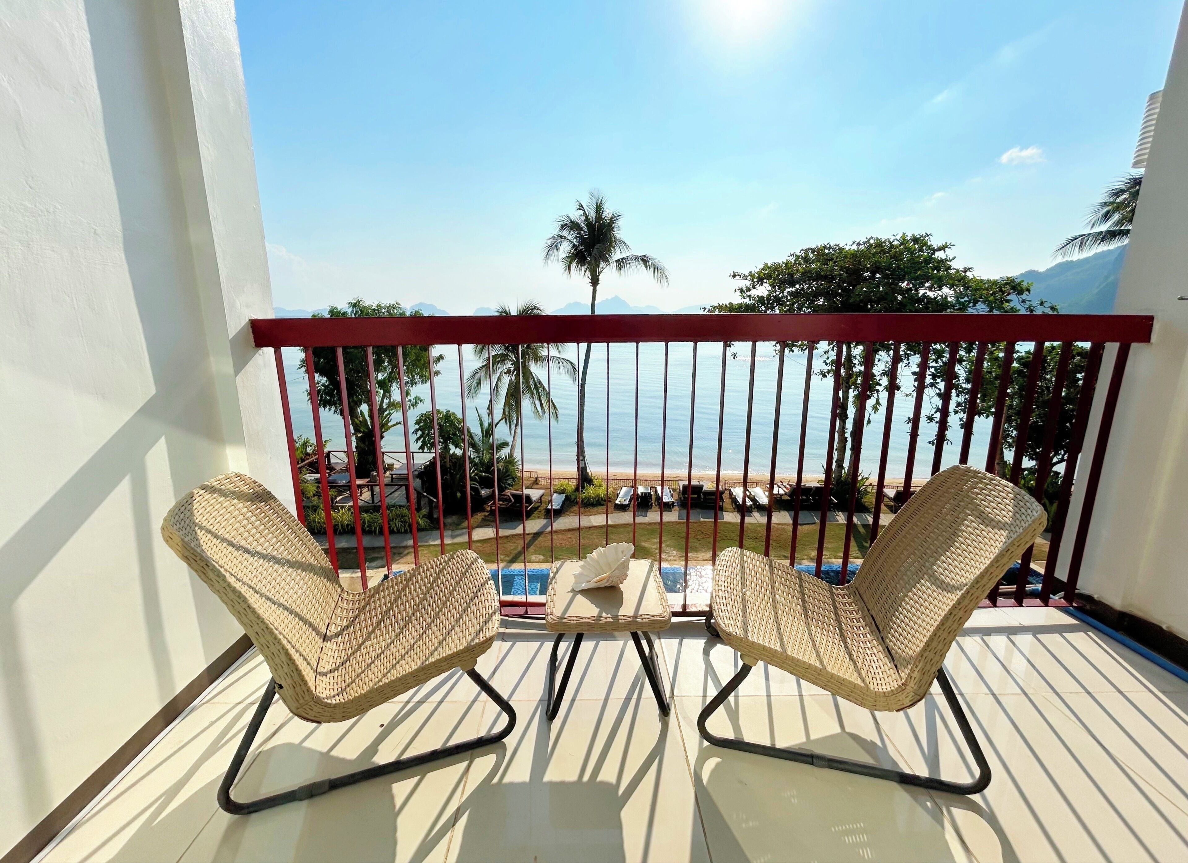 A balcony with two wicker chairs, a small table, and a view of the beach and palm trees.