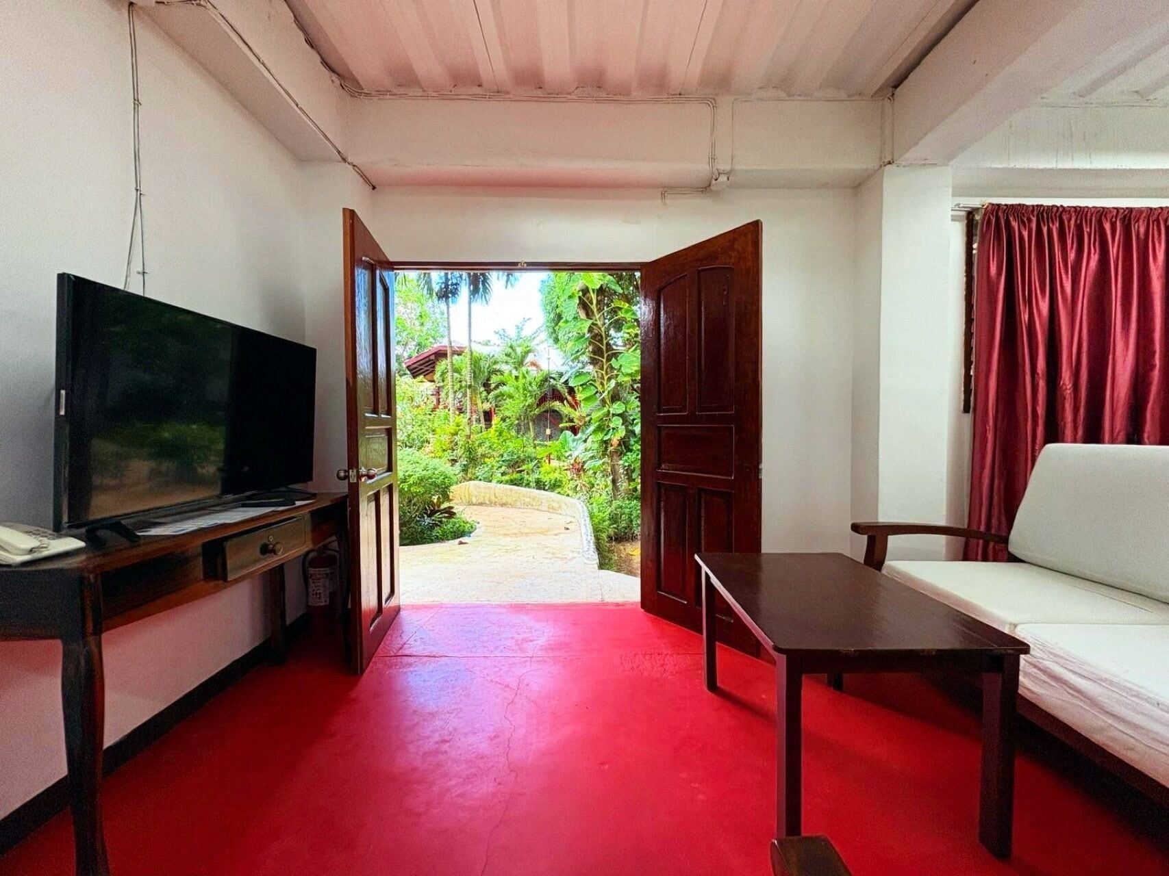A room with a TV, a table, a sofa, and a view of a garden through an open door.