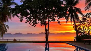 Piscina con vista al atardecer, palmeras y un barco a lo lejos.