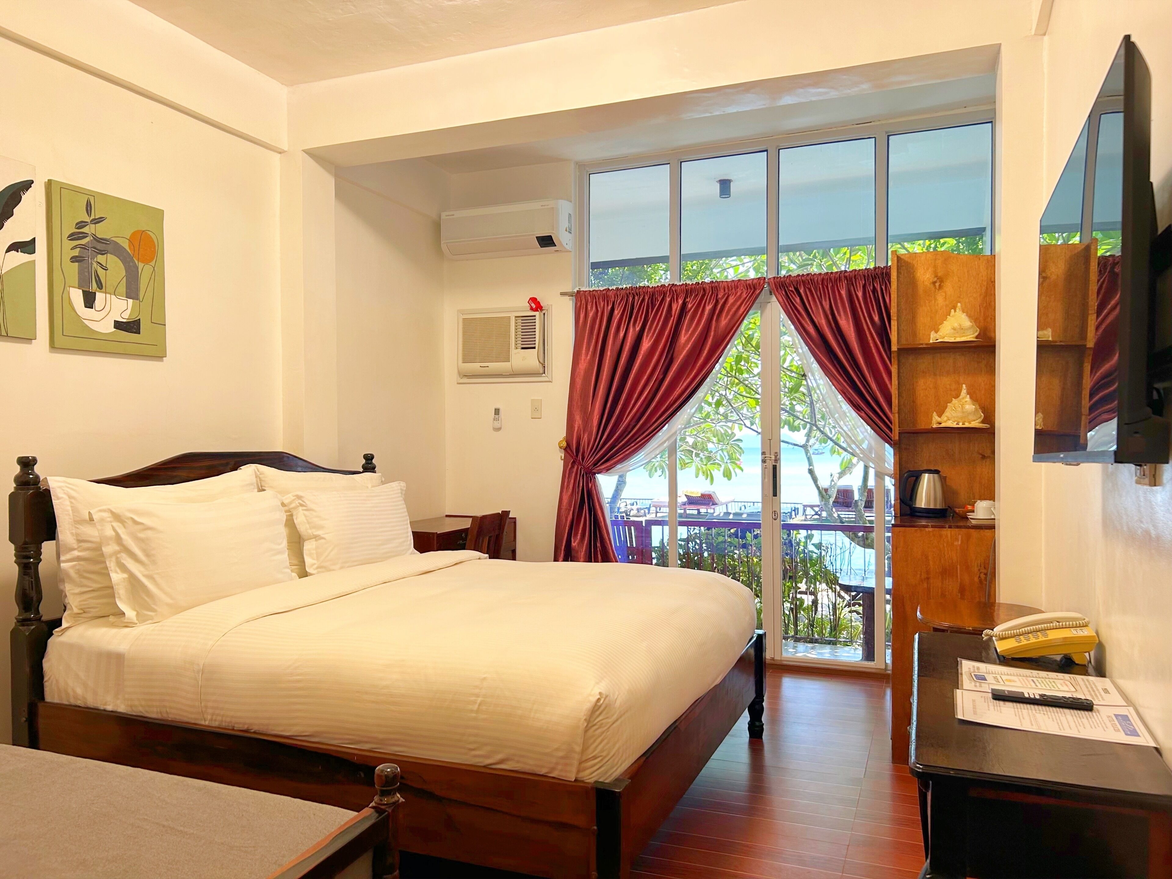 A hotel room with a bed, a desk, a chair, a TV, a window with curtains, and a balcony with a view.
