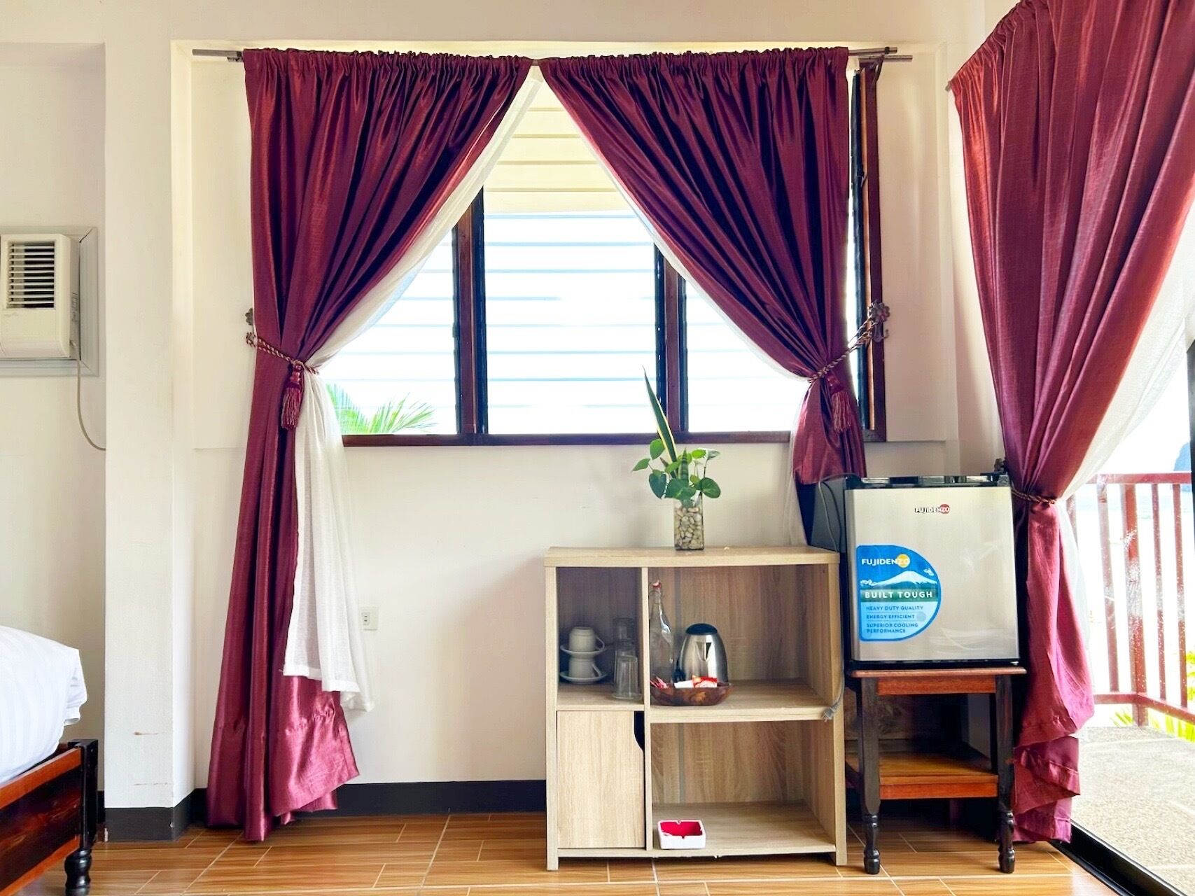 Una stanza con un letto, un piccolo mobiletto in legno con una pianta, un bollitore e un frigorifero.