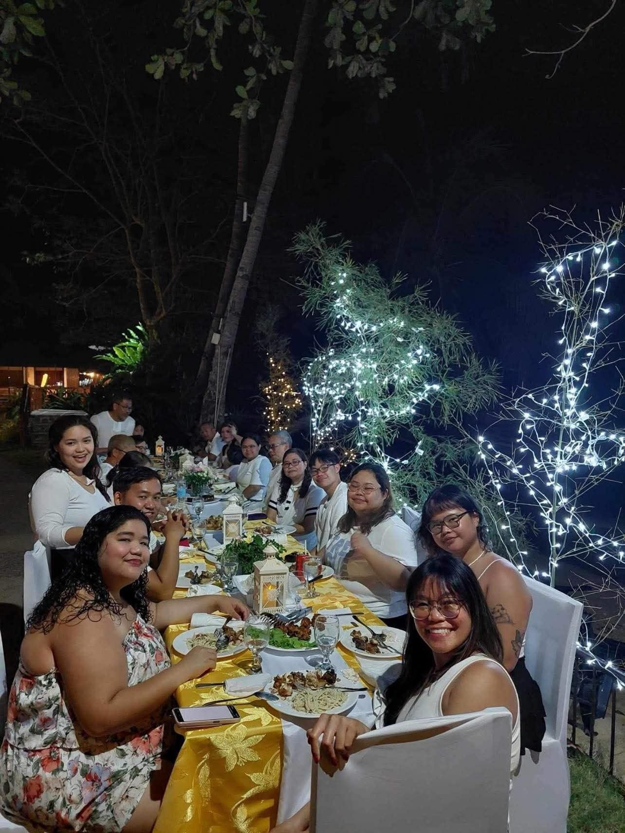 Um grupo de pessoas jantando ao ar livre à noite, iluminado por luzes de festa e com árvores ao fundo.