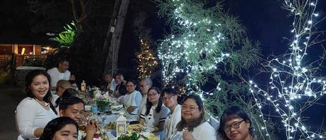 Un grupo de personas cenando al aire libre de noche, con luces colgantes y árboles al fondo.