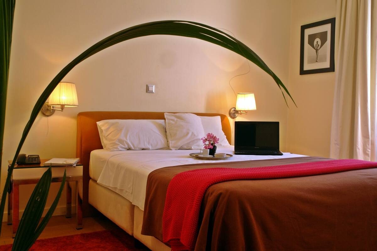 Une chambre d’hôtel avec un lit, des lampes de chevet, un bureau avec un téléphone, une petite table avec un vase de fleurs et un tableau au mur.