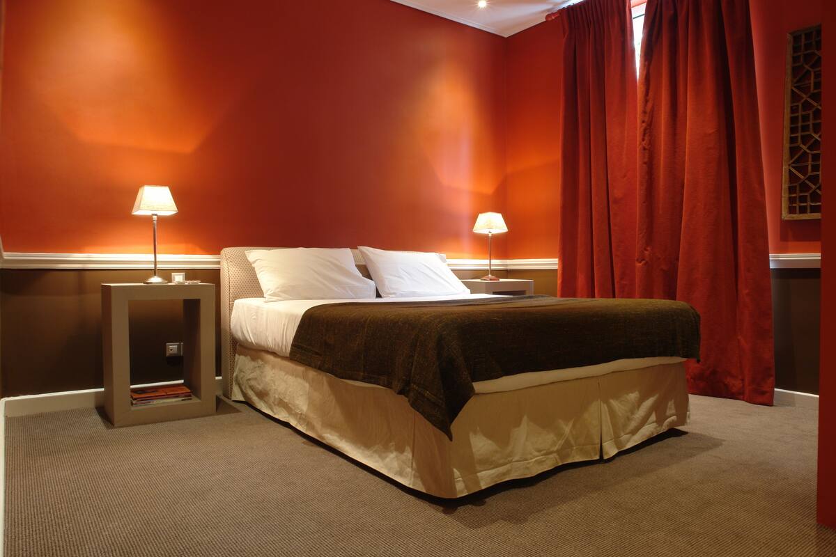 Une chambre d’hôtel avec un lit, deux lampes de chevet, une table de nuit et une fenêtre avec des rideaux rouges.