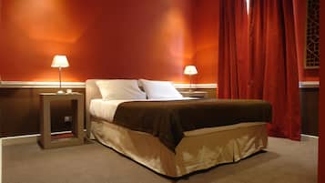 A hotel room with a bed, two bedside lamps, a nightstand, and a window with red curtains.