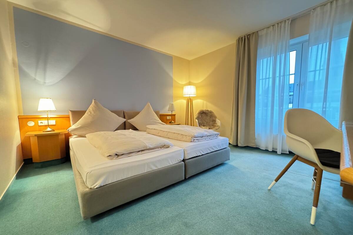 A hotel room with a double bed, two bedside lamps, a fluffy chair, and a large window with curtains.