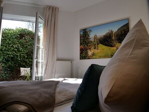 A bedroom with a bed, a window with curtains, a painting of a landscape, and a view of greenery outside. - Privathotel Bremer (Bergisch Gladbach)
