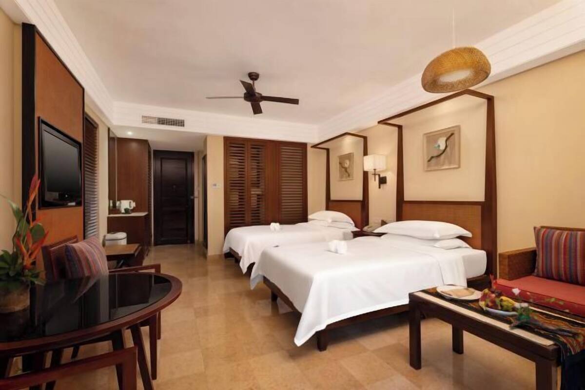 A hotel room with two beds, a dining table, a sofa, a ceiling fan, and a TV.