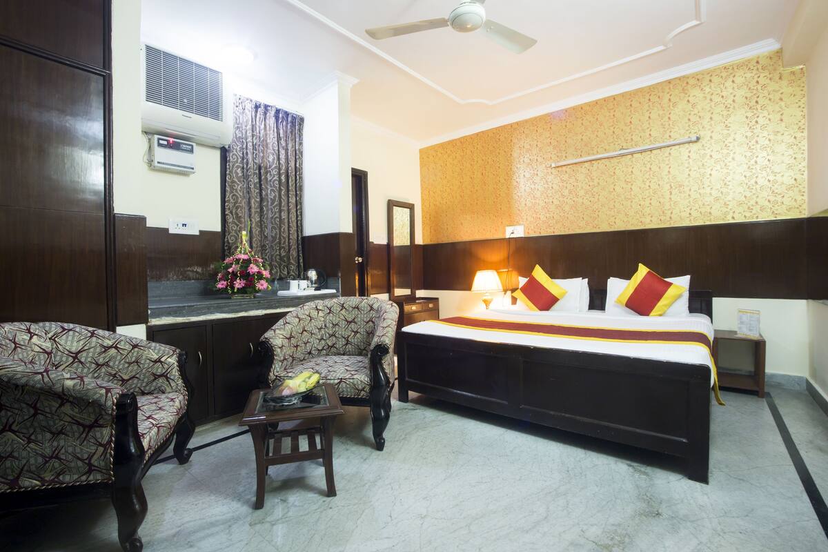 Een hotelkamer met een bed, twee fauteuils, een bijzettafeltje, een nachtkastje, een plafondventilator, een airconditioner en een decoratieve plant.