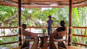 A covered outdoor seating area with wicker furniture and a central table, surrounded by lush greenery. - Cashew Grove Beach Resort (Busuanga)