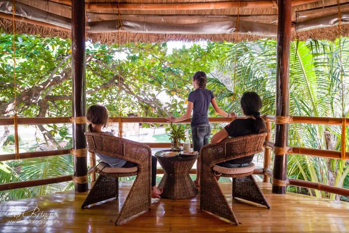 Un espacio de estar al aire libre con techo, con mobiliario de mimbre y una mesa central, rodeado de abundante vegetación.