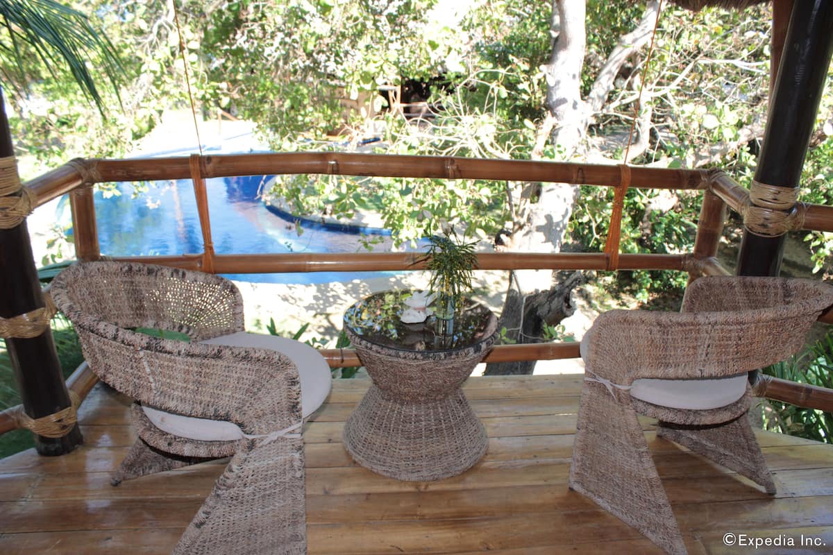 Terraza de madera con sillas de mimbre y una pequeña mesa, con vista a una piscina y abundante follaje.