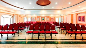 Un salón de conferencias con filas de sillas rojas, una larga mesa y un gran emblema circular en la pared.