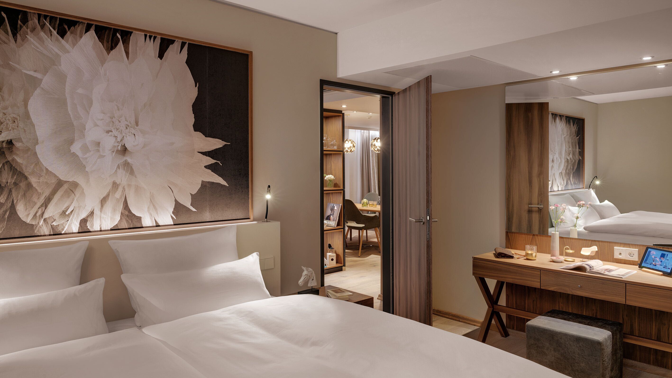 Habitació d'hotel amb un llit gran, un escriptori amb un ordinador, un mirall i una zona de menjador visible a través d'una porta oberta.