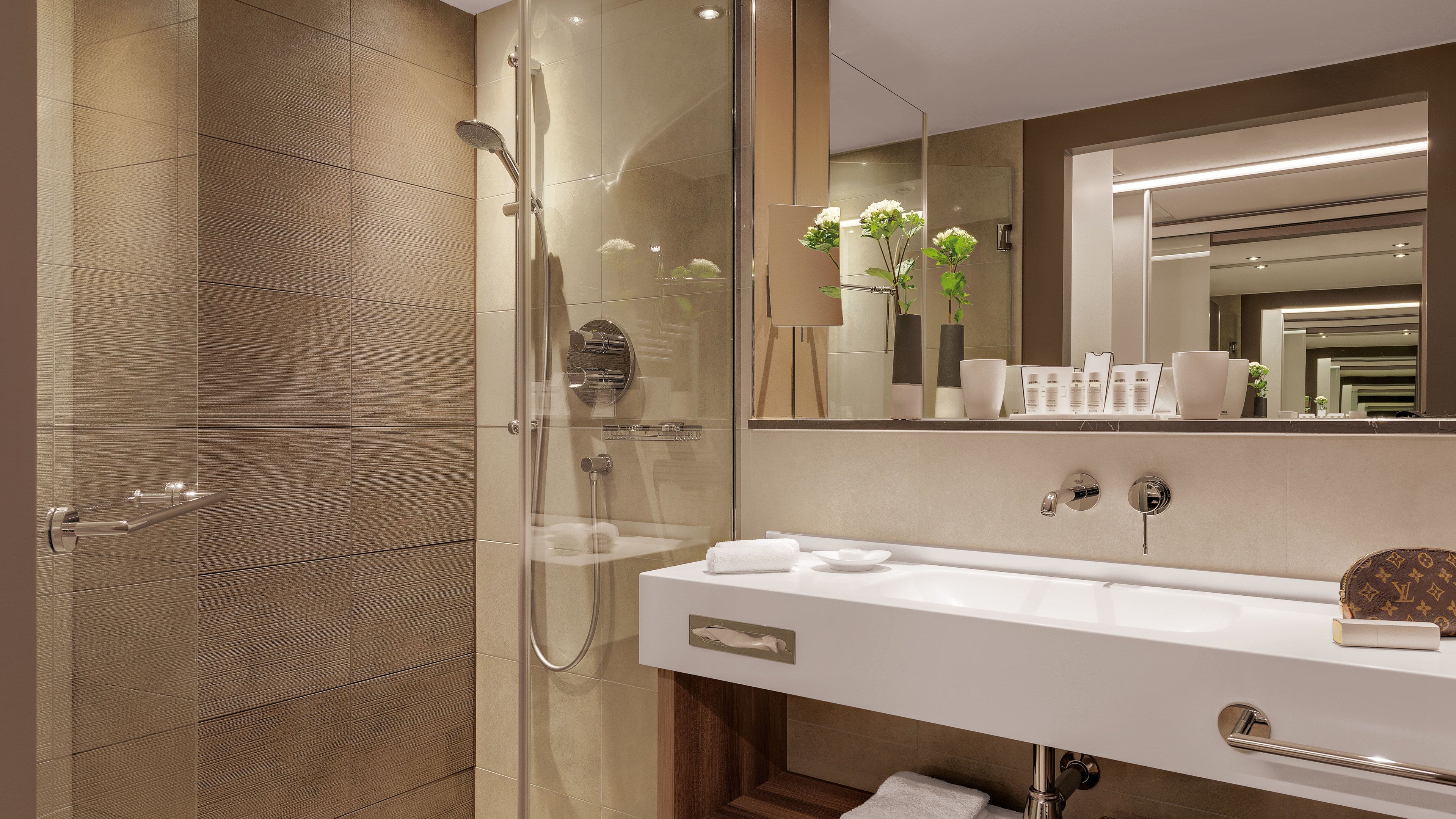 Un baño moderno con cabina de ducha de vidrio, tocador blanco con un espejo grande y un bolso Louis Vuitton sobre la encimera.