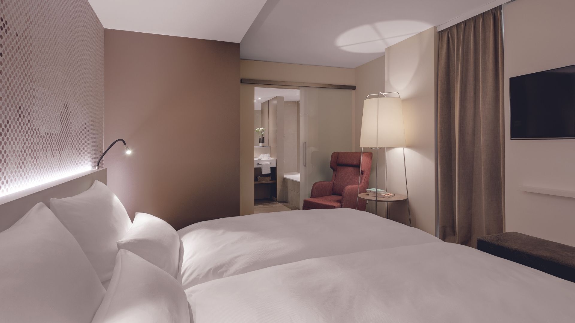 Ein Hotelzimmer mit einem großen Bett, einem Sitzbereich mit Sessel und Tisch, einem Fernseher und einem durch eine offene Tür einsehbaren Badezimmer.