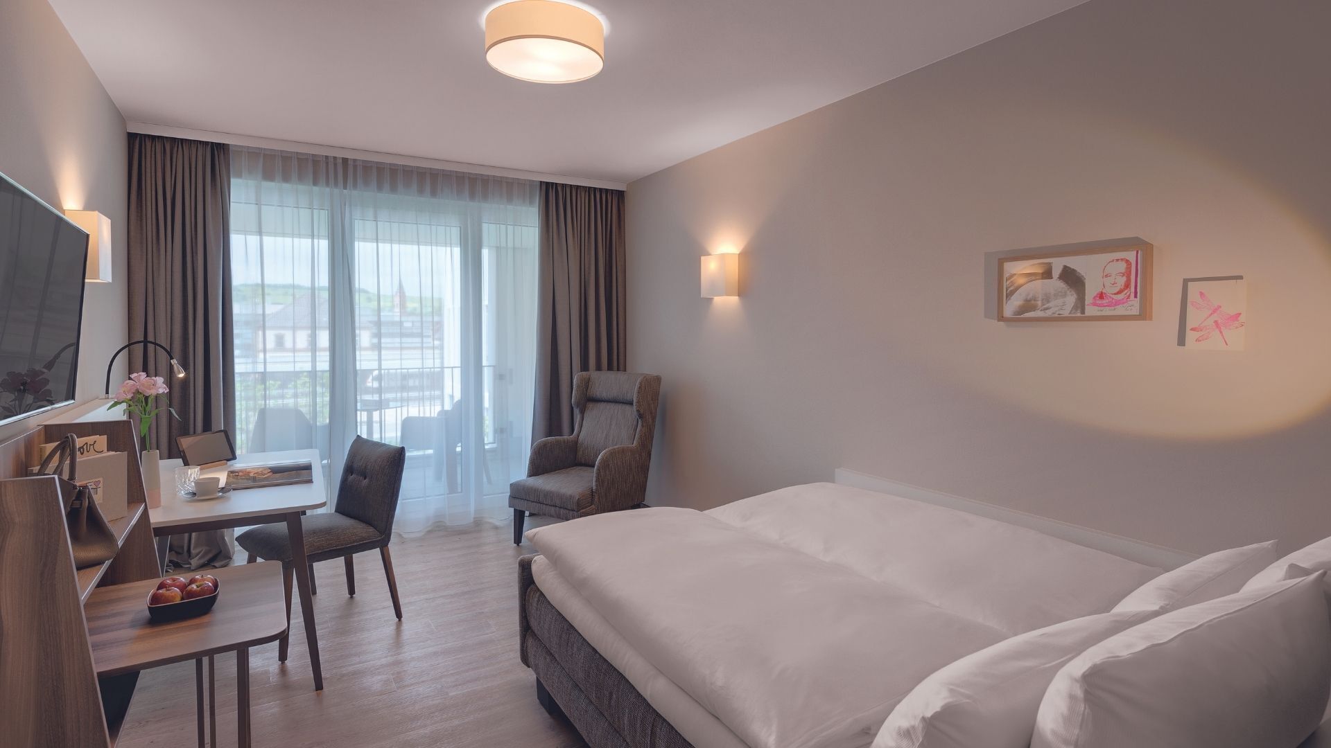 A hotel room with a bed, a desk with a laptop, a chair, a TV, and a window with curtains.