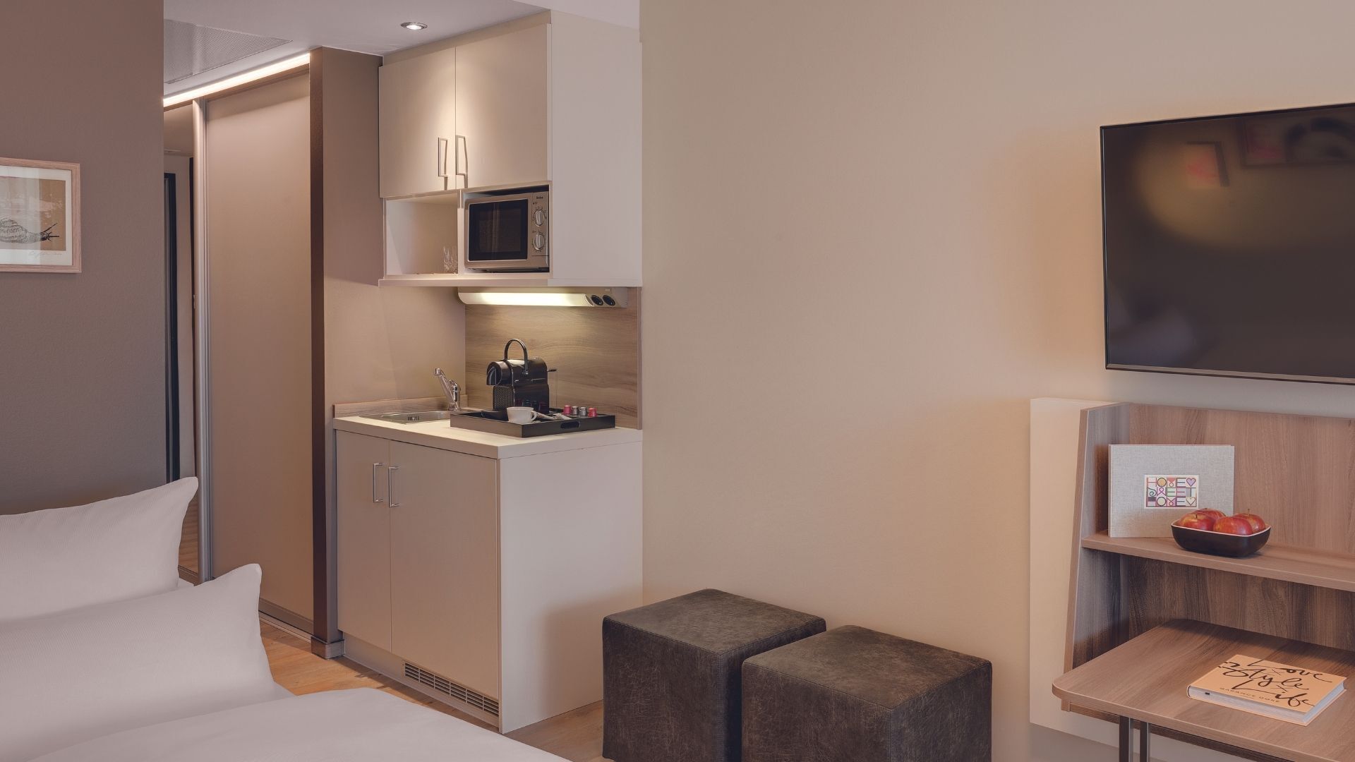 A modern hotel room with a kitchenette, a flat-screen TV, a bedside table with a book, and a small seating area.