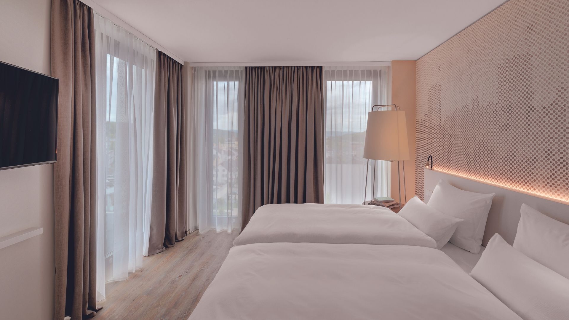 Habitación de hotel con dos camas, televisión, lámpara y vistas a la ciudad a través de amplias ventanas.