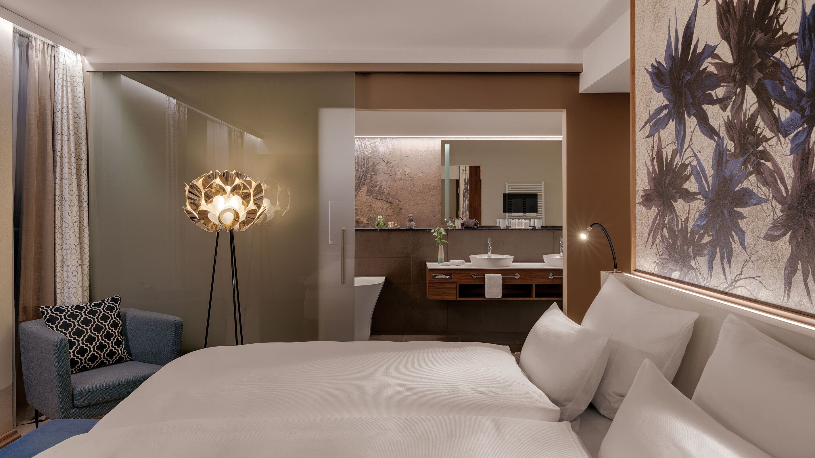 A hotel room with a large bed, a bedside lamp, a sofa, a bathroom with a sink and mirror, and a decorative wall piece.