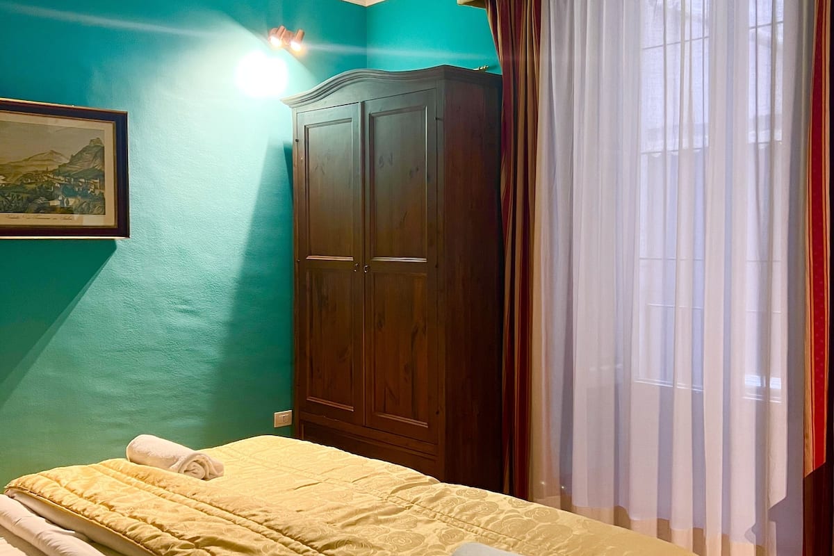 A hotel room with a large bed, a wooden wardrobe, a painting on the wall, and a window with curtains.