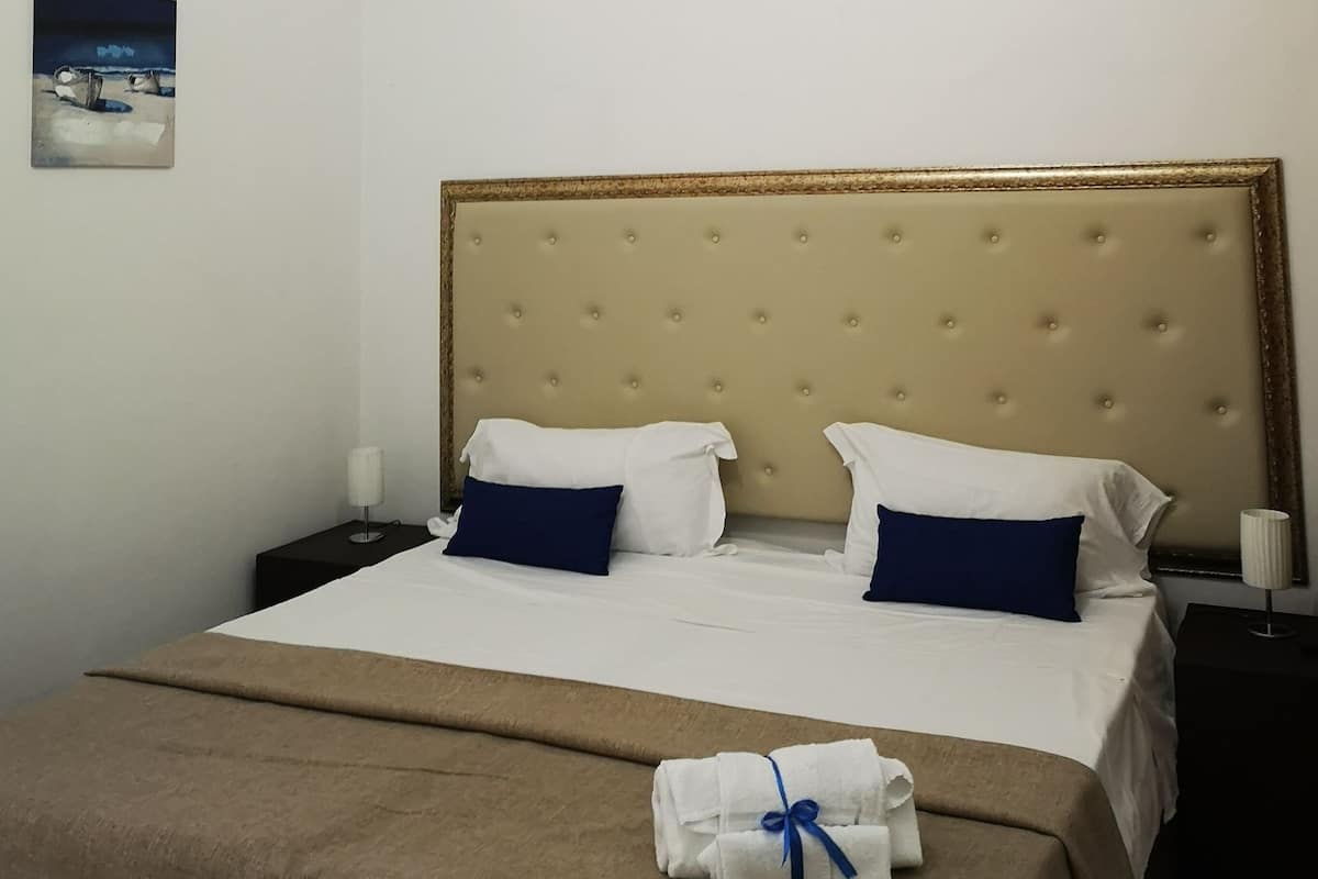 A neatly made bed with a beige headboard, white and blue pillows, and a brown bedspread. There is a small painting on the wall and a lamp on the bedside table.