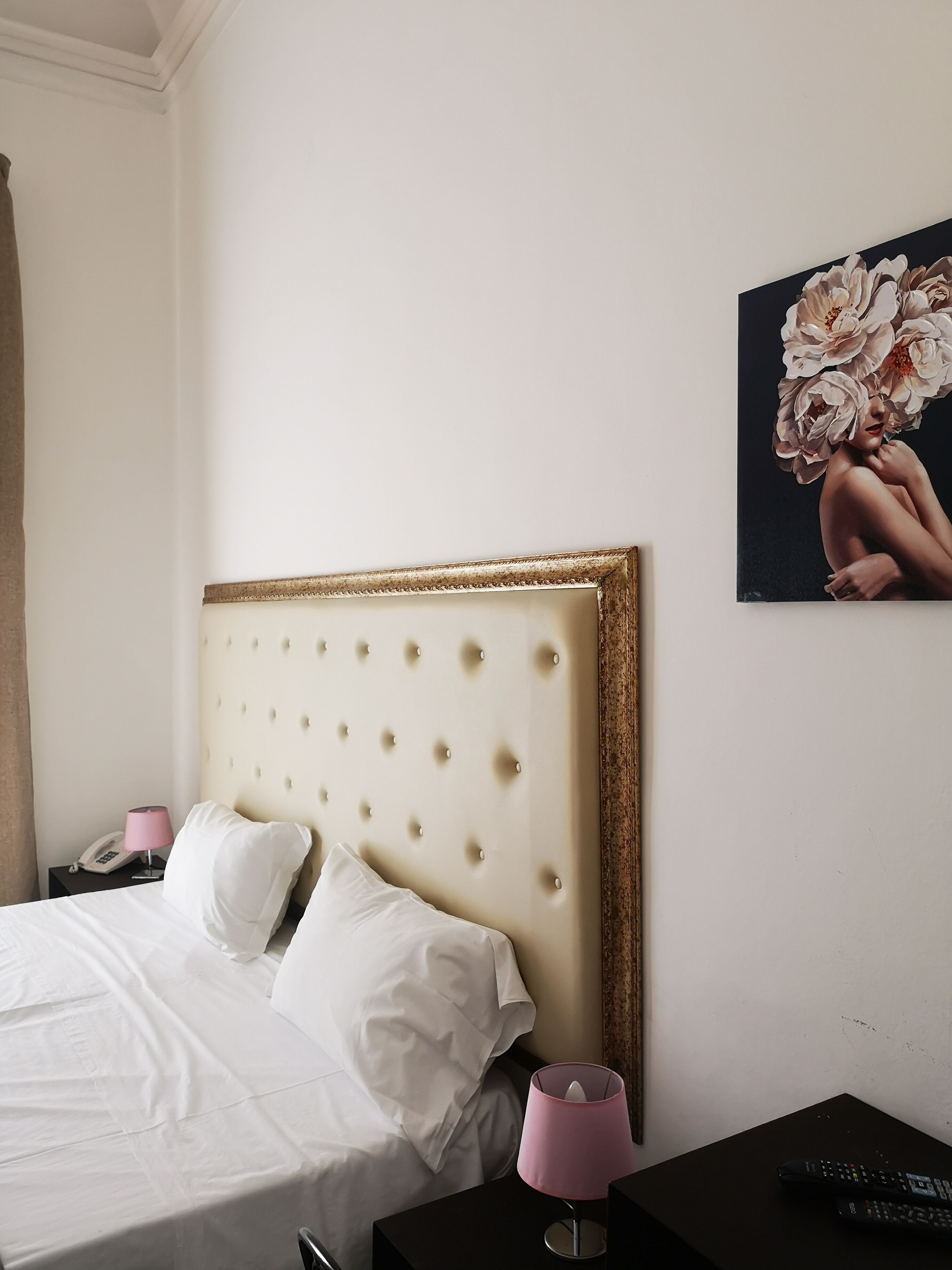 Camera d'albergo con una testiera trapuntata, biancheria da letto bianca, una lampada da comodino, un telefono e un quadro con fiori sulla parete.