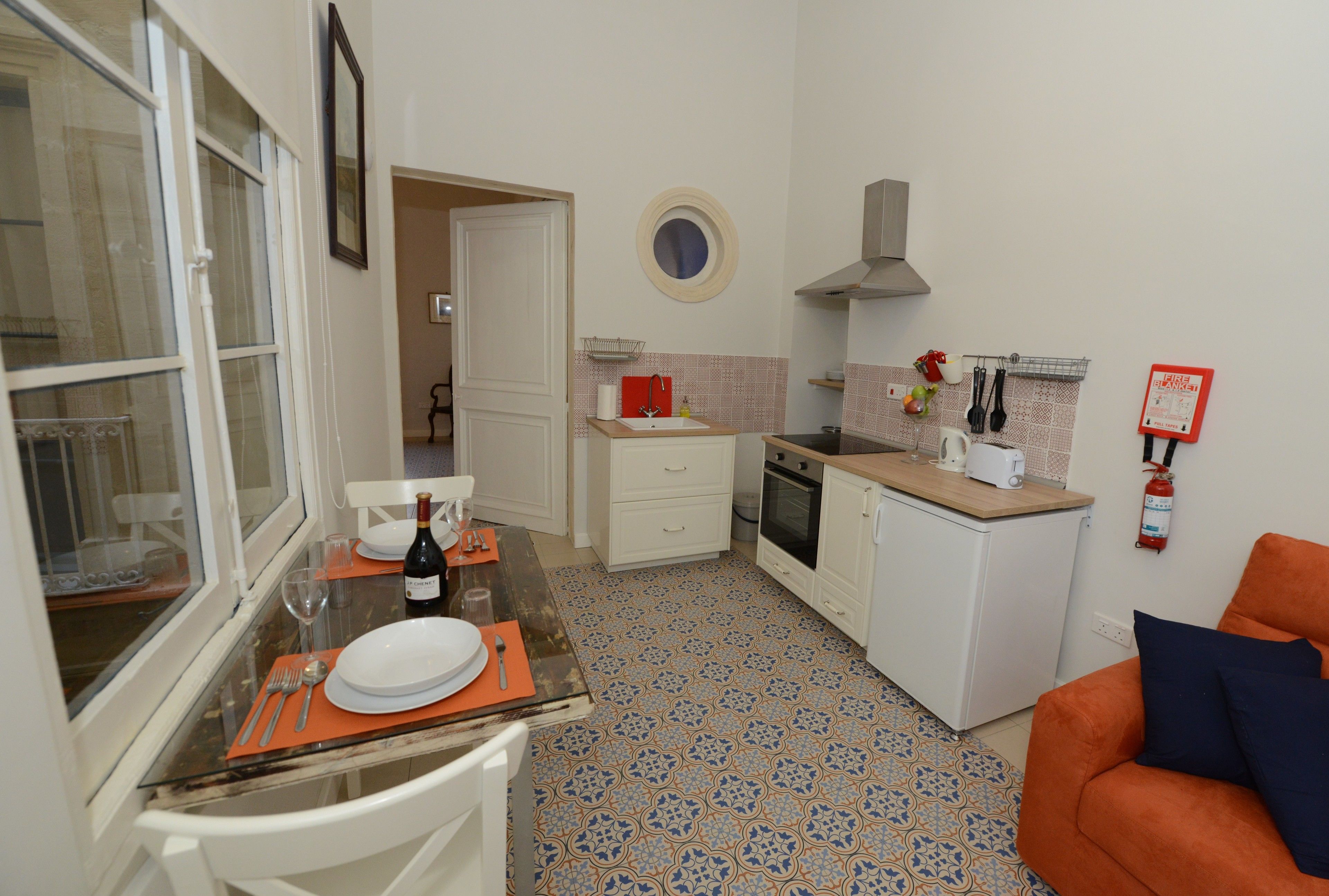 A compact kitchen with white cabinets, a small dining area with a table set for two, and a cozy orange couch in the living area.