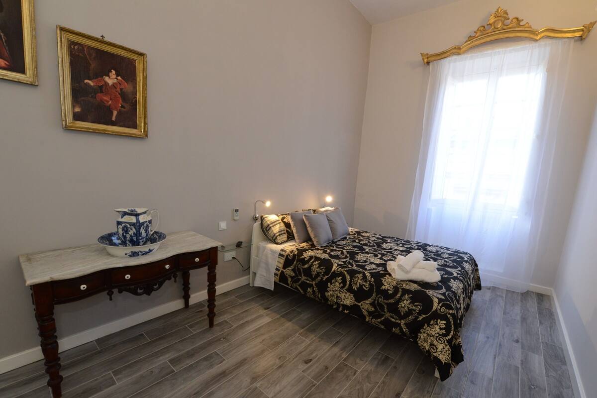 A bedroom with a bed, a bedside table, a blue and white teapot, and two framed pictures on the wall.