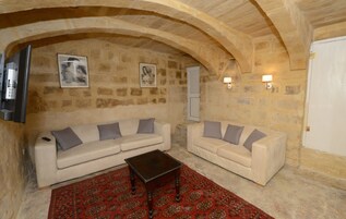 Una sala de estar acogedora con dos sofás blancos, una mesa de centro de madera oscura y un tapete rojo estampado. Se observan paredes de piedra y una puerta blanca.