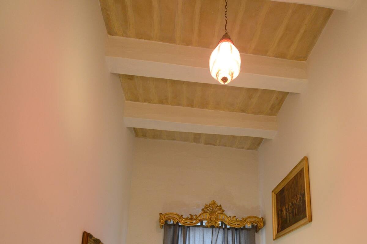 A bedroom with a bed, a nightstand, a hanging light fixture, a window with curtains, and a framed picture on the wall.