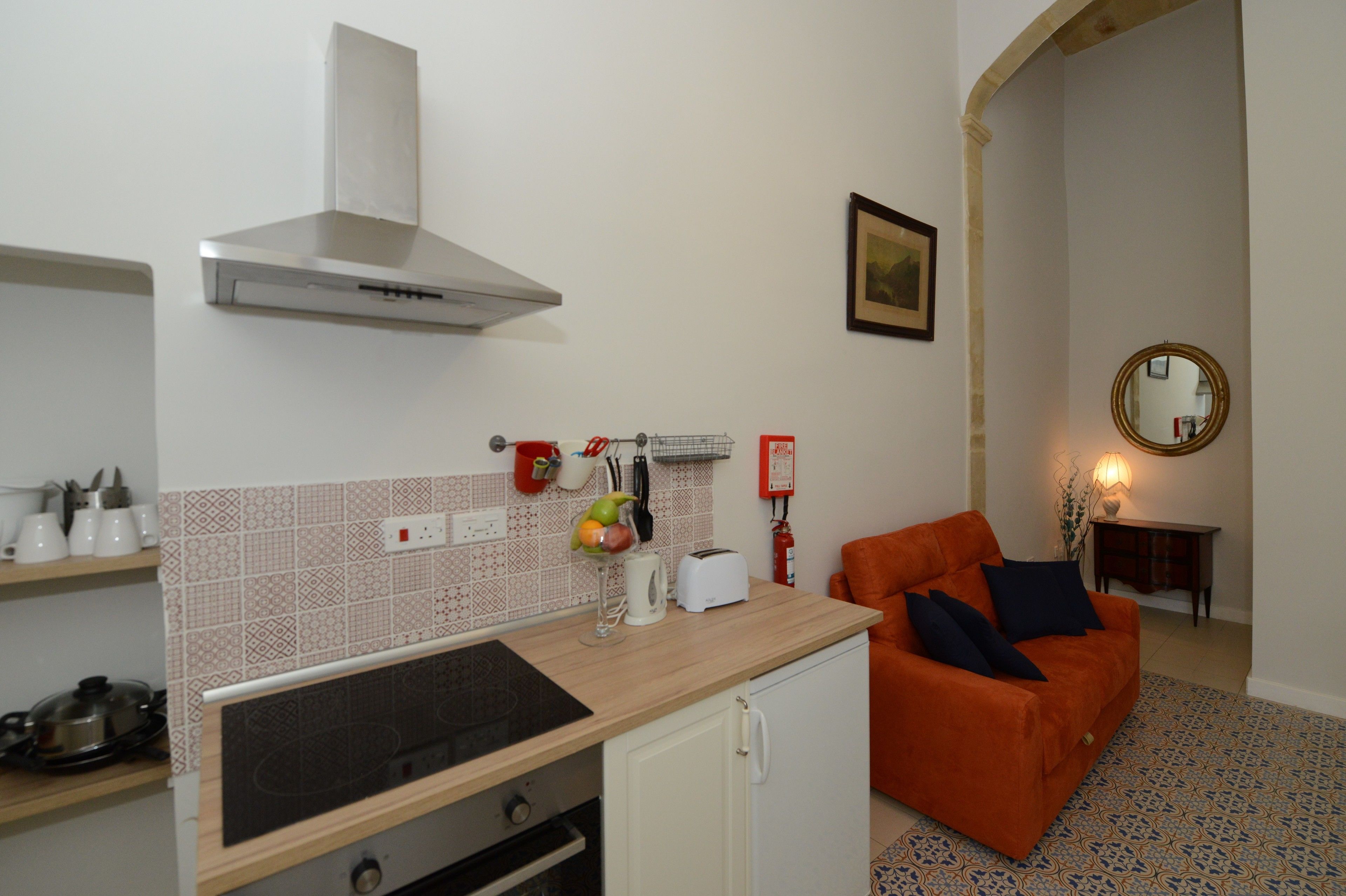 A compact kitchen with a stove, toaster, and a small cabinet. There is a red chair and a mirror in the adjacent living area.