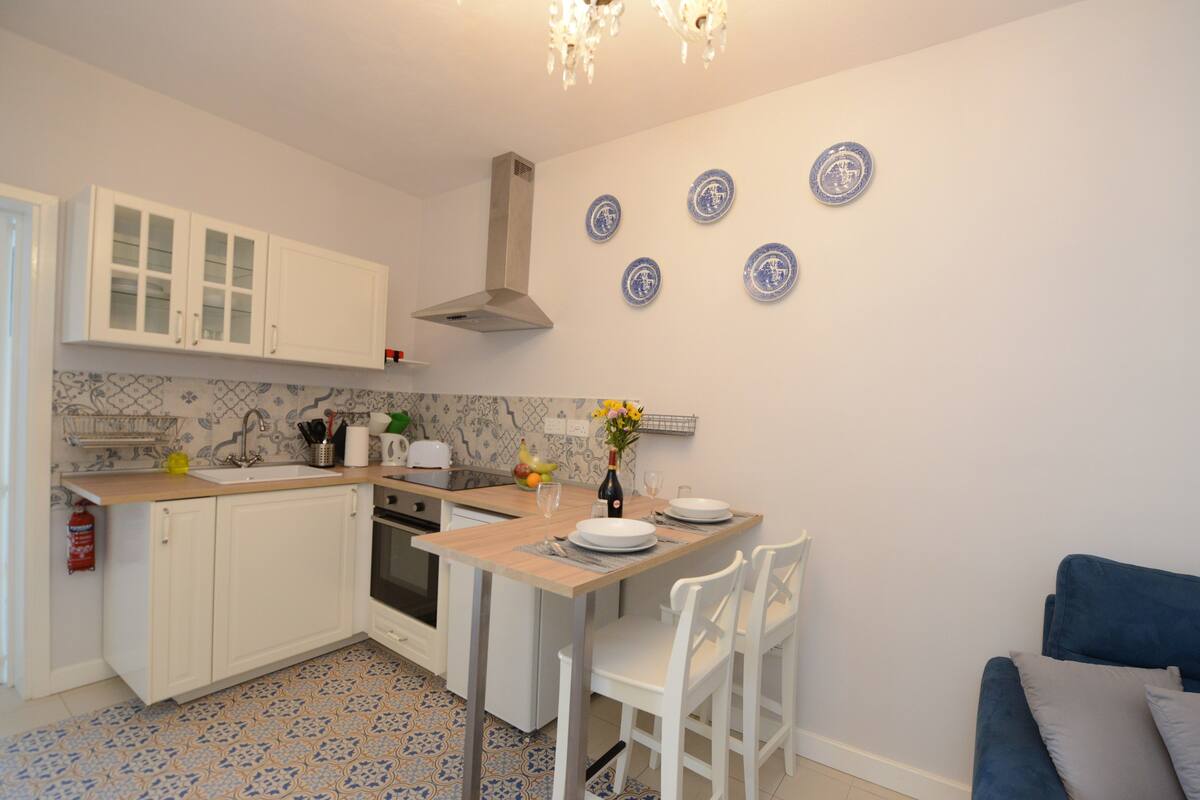 A kitchen with white cabinets, a wooden countertop, and a dining area with white chairs. There are blue plates on the wall and a vase with flowers on the counter.