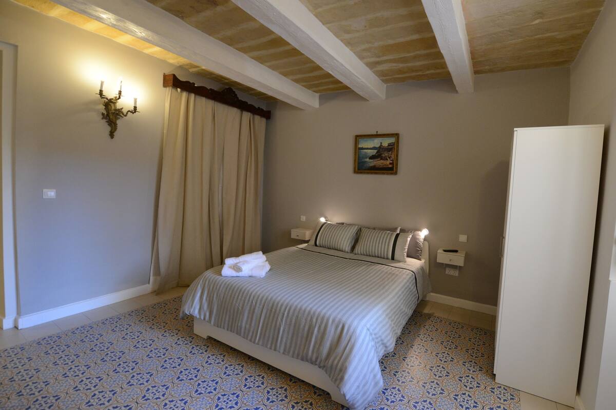 A bedroom with a bed, a nightstand, a wall-mounted lamp, a painting, and a refrigerator.