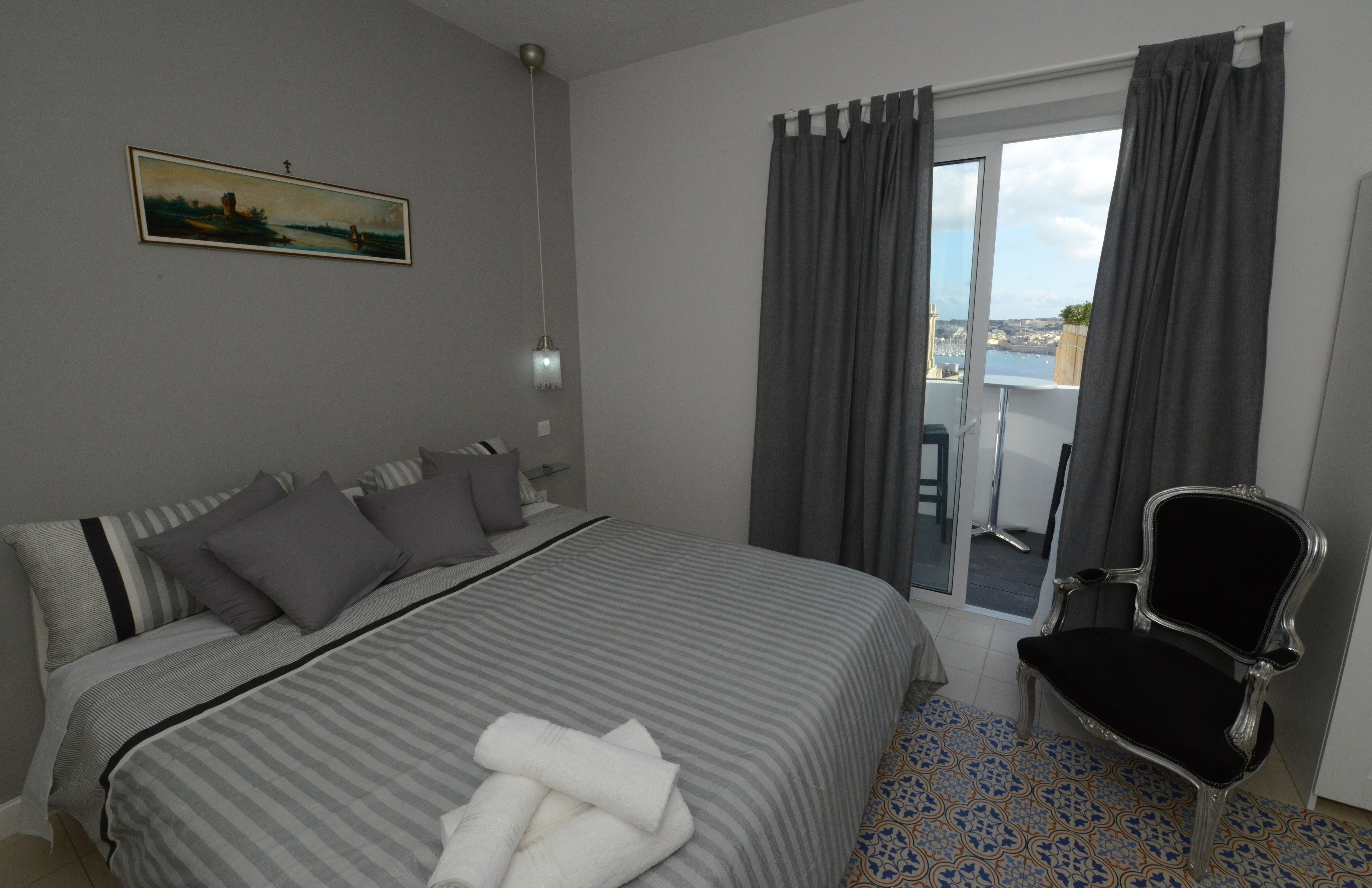 A hotel room with a bed, a chair, a painting on the wall, and a view through the window.