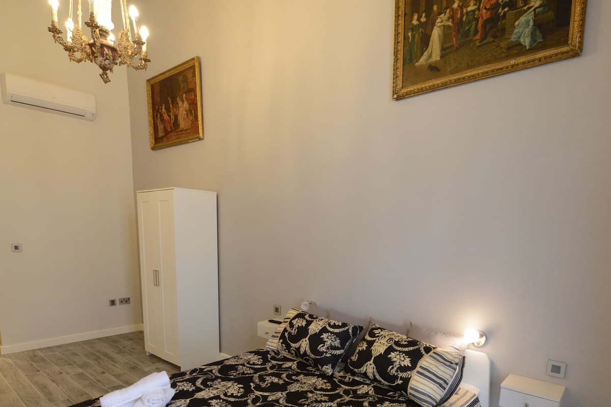 A bedroom with a bed, a chandelier, a painting, a wardrobe, and a bedside table.